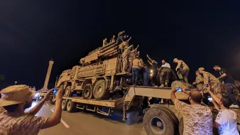 L'image montre un groupe de personnes rassemblées autour d'un véhicule militaire sur une route, probablement pendant la nuit. Plusieurs individus sont montés sur le véhicule et semblent exprimer des émotions fortes, peut-être en célébrant ou en manifestant un soutien. D'autres personnes prennent des photos avec leurs téléphones. L'ambiance générale semble dynamique et impliquée, avec une forte présence militaire sur le site.