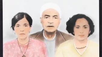 L'image représente un portrait de famille en couleur, où l'on voit trois personnes. Au centre se trouve un homme âgé avec une barbe grise, portant un chapeau blanc et un vêtement sobre. De chaque côté de lui, il y a deux femmes. À gauche, une femme avec des cheveux sombres, portant un haut rose et un collier. À droite, une autre femme avec des cheveux bouclés, habillée d'un haut jaune, qui affiche également un collier. L'arrière-plan est lumineux et flou, mettant en valeur les visages des sujets.
