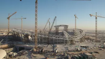 L'image montre un grand chantier de construction d'un stade, avec plusieurs grues en fonctionnement. Le stade a une structure moderne et architecturale, avec des éléments en acier visibles. En arrière-plan, on peut voir un ciel clair, suggérant qu'il fait jour. Le site est entouré de divers matériaux de construction et d'équipements, ce qui indique que le projet est encore en cours.