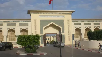 L'image montre l'entrée d'un bâtiment impressionnant, probablement une institution ou un musée, avec une architecture marocaine caractéristique. On peut voir une grande porte ornée de motifs traditionnels et des murs décorés avec des inscriptions en arabe et en français. Au-dessus de l'entrée, un drapeau rouge flotte. L'environnement est urbain, avec quelques arbres et des voitures stationnées. Le ciel est partiellement nuageux, apportant une atmosphère calme.