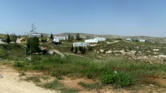 L'image montre un paysage rural avec des collines à l'arrière-plan. On peut voir plusieurs maisons en métal ou modulaires, disposées de manière dispersée. La végétation est sparse, avec des zones de terre et de l'herbe verte. Le ciel est clair et ensoleillé. L'ensemble donne une impression d'isolement et de simplicité.