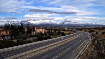 L'image montre une route large et bien entretenue, qui s'étend à travers un paysage pittoresque. On peut voir des montagnes majestueuses au loin, avec des sommets enneigés qui contrastent avec le ciel bleu parsemé de nuages. De chaque côté de la route, il y a quelques bâtiments et de la végétation, ce qui ajoute à la beauté de la scène. Cette vue évoque une ambiance paisible et naturelle, avec un mélange de modernité et de paysages ruraux.