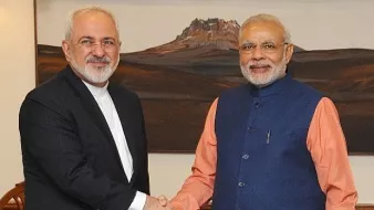 L'image montre deux hommes se serrant la main dans un cadre formel. L'un d'eux porte un costume noir et une chemise blanche, tandis que l'autre est habillé d'une veste bleue et d'une chemise orange. Ils semblent être en train d'échanger une salutation amicale, et l'arrière-plan montre une œuvre d'art représentant un paysage. L'atmosphère semble cordial et professionnelle.