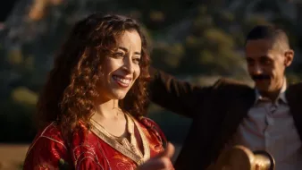 L'image montre une femme souriante avec des cheveux bouclés, portant une robe rouge ornée de motifs. Elle semble joyeuse et est entourée d'un environnement naturel. Un homme en arrière-plan, vêtu d'un costume, semble également profiter du moment. L'atmosphère générale est festive et conviviale.