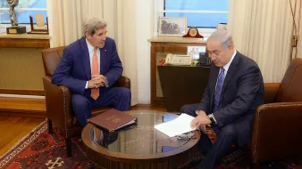 L'image montre deux hommes assis dans une pièce restructurée. L'un est vêtu d'un costume orange et l'autre d'un costume sombre, tous deux semblent être en discussion sérieuse. Ils sont assis autour d'une table basse en bois, avec des documents devant eux. En arrière-plan, on aperçoit une fenêtre et une décoration sobre, suggérant un cadre officiel et professionnel.