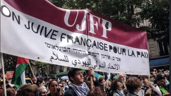 L'image montre une manifestation où des personnes tiennent une grande banderole. Cette banderole mentionne "UJFP" et affiche des messages en français, hébreu et arabe, qui semblent appeler à la paix. Le contexte suggère un rassemblement pour des causes sociales ou politiques, probablement en lien avec la paix au Moyen-Orient. En arrière-plan, on voit une foule de manifestants, des arbres et des bâtiments, ce qui indique que l'événement se déroule en milieu urbain.