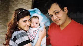 Sur cette image, on voit un couple et un bébé. La femme, à gauche, sourit et tient le bébé enveloppé dans une couverture, tandis qu'un homme, à droite, regarde la caméra en souriant également. Ils se tiennent tous les trois dans un environnement intérieur, probablement chez eux, avec un mur en briques en arrière-plan. L'atmosphère générale est chaleureuse et affectueuse.