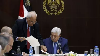 L'image montre une réunion, probablement politique, avec plusieurs hommes assis à une table. Au premier plan, un homme, probablement un leader ou un officiel, est assis regardant des documents. Un autre homme, debout à ses côtés, semble lui présenter des papiers ou des informations. En arrière-plan, on peut voir un drapeau, ainsi qu'un emblème qui semble représenter une organisation ou un gouvernement. L'ambiance générale de la scène est sérieuse et attentive, suggérant une discussion importante.