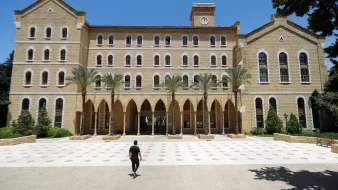 L'image montre un grand bâtiment en pierre avec une architecture traditionnelle, comportant plusieurs fenêtres à arc et des colonnes. Au premier plan, une personne marche seule sur une place pavée, tandis que des palmiers sont visibles sur les côtés. Le ciel est clair et bleu, ce qui donne une atmosphère ensoleillée. Le bâtiment semble être une institution éducative ou un lieu historique.