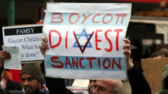 L'image montre des manifestants tenant des pancartes lors d'une manifestation. L'une des pancartes, bien visible, porte les mots "BOYCOTT, DIVEST, SANCTION" avec un symbole en forme d'étoile. Les manifestants semblent exprimés des opinions politiques ou sociales fortes, et l'ambiance de l'événement semble engagée.