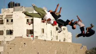 L'image montre un groupe de jeunes en train de sauter par-dessus un mur. Ils semblent s'amuser à réaliser des acrobaties, témoignant d'une grande énergie et d'une certaine audace. En arrière-plan, on aperçoit des bâtiments avec des murs marqués, ce qui donne une idée de l'environnement urbain. Le ciel est clair, ce qui contraste avec les structures des bâtiments. L'ambiance générale de l'image est dynamique et vivante, capturant un moment de loisirs et de camaraderie entre les adolescents.