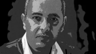 L'image montre un homme avec une expression réfléchie. Il a les cheveux courts et porte un pull. Ses mains sont croisées devant lui. Le fond est sombre, ce qui met en avant son visage. L'éclairage accentue ses traits et donne une atmosphère sérieuse à la photo.