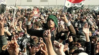 L'image montre une grande foule rassemblée, probablement lors d'une manifestation ou d'un événement public. Les personnes levées les bras, brandissant des drapeaux, affichent des expressions d'énergie et d'engagement. Certains portent des vêtements traditionnels, et l'ambiance semble dynamique et fervente. Des pancartes ou des symboles visibles pourraient suggérer un message politique ou social. L'environnement semble être un espace ouvert, peut-être une place ou une route.