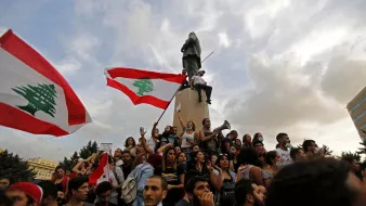 L'image montre une manifestation au Liban. Des groupes de personnes sont rassemblés autour d'un monument, tenant des drapeaux libanais. Certains manifestants chantent ou s'expriment à l'aide de mégaphones. L'atmosphère semble dynamique et engagée, avec des nuages dans le ciel qui ajoutent une ambiance dramatique à la scène.