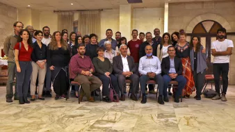 L'image montre un grand groupe de personnes rassemblées dans une salle, probablement pour un événement ou une célébration. Les gens sont assis et debout, souriant et regardant vers l'objectif. L'espace présente des murs en pierre et un sol en marbre, créant une atmosphère conviviale. On peut remarquer une variété de styles vestimentaires parmi les participants, ce qui indique une diversité au sein du groupe.