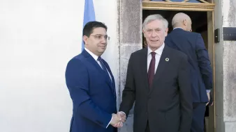 L'image montre deux hommes en train de se serrer la main, échangeant probablement une salutation ou un accord. L'un est habillé d'un costume bleu, tandis que l'autre porte un costume sombre avec une cravate. On aperçoit un drapeau derrière eux, suggérant un contexte officiel ou diplomatique. Leur expression semble amicale et professionnelle.