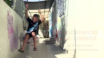 L'image montre un jeune garçon qui se balançe sur une balançoire improvisée. Il semble joyeux et se trouve dans un espace qui semble être un couloir ou une cour. Les murs autour de lui sont décorés avec des peintures murales, dont une représente un chat. Le cadre donne une impression de vie quotidienne dans un endroit où l'art et le jeu se côtoient. On voit également un message en arrière-plan, lié à des thèmes de liberté.