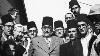 L'image représente un groupe d'hommes en costumes formels, majoritairement portant des fez. Ils semblent être engagés dans une discussion ou un moment de camaraderie. L'ambiance est sérieuse, et les expressions des visages montrent une concentration ou une détermination. Une femme est également visible en arrière-plan, portant un foulard. Le cadre pourrait indiquer un événement historique ou un rassemblement important, probablement dans un contexte culturel ou politique.