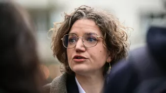 L'image montre une femme avec des cheveux bouclés et des lunettes, en train de parler. Elle semble engagée dans une conversation, peut-être lors d'un événement public. Son expression faciale est sérieuse, et elle porte un vêtement avec un col blanc et une veste. L'arrière-plan paraît flou, suggérant qu'elle se trouve au milieu d'un groupe ou d'une foule.