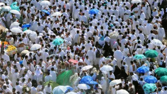 L'image montre une grande foule de personnes vêtues de vêtements blancs, probablement en train de participer à un pèlerinage ou à un événement religieux. De nombreux participants arborent des parapluies colorés, qui offrent de l'ombre dans un environnement ensoleillé. L'atmosphère semble très animée, avec un grand nombre de personnes rassemblées, ce qui reflète un moment de rassemblement spirituel.
