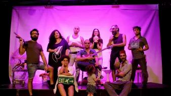 L'image montre un groupe de personnes posant sur scène dans une ambiance colorée, avec un éclairage rose. Chaque membre a une pose ou un accessoire distinct, créant un effet visuel dynamique. Certains tiennent des objets variés, comme un instrument de musique, un panneau de sortie et un appareil électronique. Au premier plan, on peut voir une petite fille debout, ajoutant une touche de légèreté à la scène. L'ensemble dégage une atmosphère artistique et joyeuse.