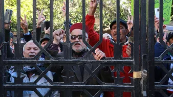 L'image montre un groupe de personnes rassemblées derrière une grille. Certains individus lèvent leurs doigts en forme de V, symbole de la paix ou de la victoire. Les expressions des participants varient, allant de la détermination à l'enthousiasme. Il semble qu'ils soient engagés dans une manifestation ou un rassemblement. Les arrière-plans sont flous, mettant l'accent sur les manifestants et leur activité.