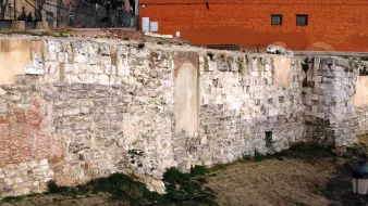 L'image montre une section de mur en pierre, probablement une ancienne structure ou fortification. Les pierres sont de tailles et de formes variées, avec des parties couvertes de végétation. En arrière-plan, on peut apercevoir un mur de couleur rouge et quelques éléments de structure moderne. Le site semble être à l'extérieur, probablement dans une zone historique, avec une ambiance de délabrement et d'abandon.