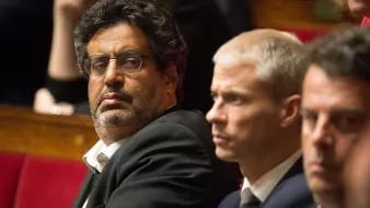 L'image montre un groupe d'hommes assis dans un cadre formel, probablement lors d'une séance législative. Un homme au premier plan porte des lunettes et un costume sombre, affichant une expression sérieuse, tandis que les autres à ses côtés semblent également attentifs et concentrés. L'arrière-plan montre des personnes floues, suggérant une ambiance politique. Les sièges sont ornés de rouge, typiques des assemblées.