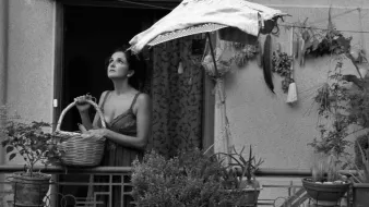L'image montre une femme se tenant sur un balcon, tenant un panier dans ses bras. Elle semble contemplative, regardant au loin. Le balcon est décoré de plusieurs plantes et herbes suspendues. Un parasol en toile protège en partie la femme du soleil. L'image est en noir et blanc, ce qui lui donne une atmosphère nostalgique et mélancolique.