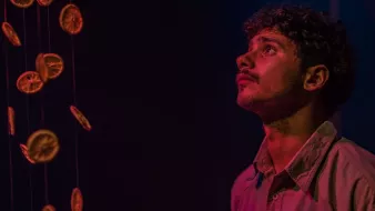 L'image montre un jeune homme regardant avec curiosité quelque chose au-dessus de lui. Il est entouré d'une ambiance sombre, avec des éclairages qui créent des reflets colorés, probablement des nuances de bleu et de rose. Des rondelles d'orange sèchées pendent de fil, créant un effet visuel intéressant, tandis que l'expression du jeune homme semble pensive et captivée par la scène.