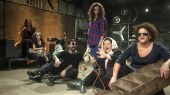 L'image présente un groupe de personnes assises sur le sol d'un espace décoratif et artistique. On y voit des femmes et des hommes, certains souriant et riant, dans une ambiance décontractée et joyeuse. Les femmes portent des vêtements variés, y compris un hijab, tandis que certains d'entre eux portent des lunettes de soleil. À l'arrière-plan, il y a des instruments de musique et des éléments de décor industriels, ce qui ajoute une touche créative à la scène. Au centre, une femme tient une chicha (narguilé) et semble engagée dans la conversation. L'ensemble de la composition donne une impression de camaraderie et de convivialité.