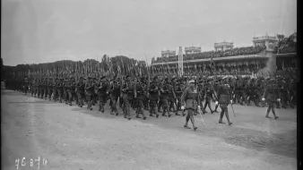 L'image montre un défilé militaire, avec de nombreux soldats marchant en formation. Ils portent des uniformes d'époque et tiennent des fusils à la main. En arrière-plan, on peut voir une tribune avec des spectateurs, probablement des dignitaires ou des spectateurs admirant le défilé. L'atmosphère semble solennelle et organisée, typique des événements militaires de ce genre. Le ciel est nuageux, ce qui donne une ambiance sobre à la scène.