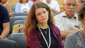 L'image montre une femme assise dans une salle de conférence ou de réunion, avec une expression pensive. Elle porte un haut de couleur sombre et a de longs cheveux bouclés. En arrière-plan, on peut voir d'autres personnes dans une atmosphère assez calme. La femme semble concentrée sur ce qui se passe autour d'elle.