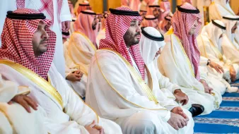 L'image montre un groupe d'hommes assis, probablement en train de prier ou de méditer. Ils portent des vêtements traditionnels saoudiens, comme des thobes blancs et des chapeaux appelés ghutras, souvent ornés de bandes rouges et blanches. L'atmosphère semble empreinte de sérénité et de respect, reflet d'un moment spirituel partagé. Le décor autour d'eux est élégant et peut inclure des éléments architecturaux typiques d'un lieu de culte.