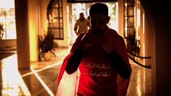 L'image montre un homme portant un t-shirt rouge avec un drapeau. Il semble s'apprêter à sortir, avec une ambiance chaleureuse et lumineuse en arrière-plan. On peut voir des éléments de décor qui suggèrent un espace intérieur, avec des silhouettes floues et une lumière dorée qui crée une atmosphère dramatique. L'homme porte une expression sérieuse et déterminée.