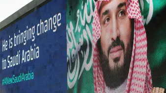 L'image montre une grande affiche murale présentant un homme en tenue traditionnelle saoudienne, avec un keffieh sur la tête. À côté de son visage, il y a un message en anglais qui dit "He is bringing change to Saudi Arabia" et un hashtag "#NewSaudiArabia". L'arrière-plan de l'affiche est bleu et le texte blanc. Cela évoque un thème de changement et de modernisation en Arabie Saoudite.