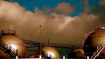 L'image présente des réservoirs à gaz de forme sphérique, surmontés de structures métalliques. Ils sont disposés sur une plateforme, tandis qu'un ciel nuageux et dramatique se profile en arrière-plan. Les couleurs à la fois sombres et lumineuses créent une atmosphère industrielle marquée par le contraste entre les réservoirs et le ciel.