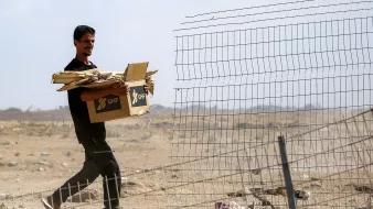 Homme portant une boîte et du bois près d'une clôture dans un paysage désertique.