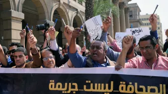 L'image montre une manifestation avec un groupe de personnes levant les bras et tenant des pancartes. Ils semblent exprimer leur mécontentement ou revendiquer des droits, notamment en rapport avec la liberté de la presse. L'atmosphère est énergique et déterminée, avec des visages montrant des émotions de passion et de revendication. À l'arrière-plan, on peut voir des bâtiments qui indiquent un environnement urbain. Le slogan sur la banderole semble défendre une position sur la presse, suggérant que celle-ci ne devrait pas être considérée comme un crime.