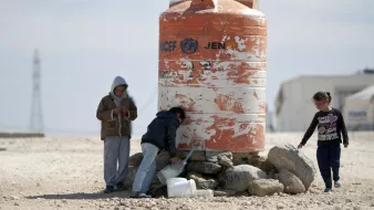 L'image montre un groupe d'enfants autour d'un réservoir d'eau. Le réservoir, de couleur orange et marqué par des signes d'usure, semble abriter l'eau potable. Les enfants, dont l'un est en train de remplir un récipient, sont habillés de manière simple et semblent se trouver dans un environnement désertique. On peut voir des pierres autour du réservoir, et le paysage en arrière-plan est sobre, suggérant une situation de besoin en ressources.