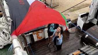 L'image montre deux personnes sur un bateau, sous un grand drapeau noir, rouge et vert. L'une des personnes sourit et pose pour la photo, tandis que l'autre semble tenir le drapeau. Le décor autour inclut du bois, des cordages et d'autres éléments d'un environnement nautique. L'atmosphère semble conviviale et dynamique.