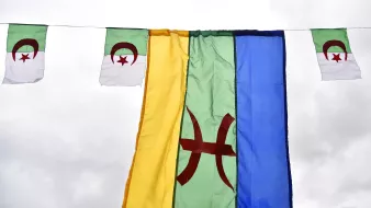 L'image montre plusieurs drapeaux suspendus. Au centre, il y a un grand drapeau de couleur verte avec un symbole rouge au centre, probablement représentant le mouvement amazigh. De chaque côté, il y a de plus petits drapeaux, qui semblent être le drapeau algérien, avec les couleurs verte et blanche et le croissant et l'étoile rouges. L'ensemble est accroché sous un ciel nuageux, créant une ambiance festive ou symbolique.