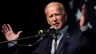 L'image montre un homme en train de donner un discours. Il est debout derrière un microphone et utilise ses mains pour gesticuler, ce qui suggère qu'il est passionné par ce qu'il dit. Il porte un costume bleu sombre avec une chemise à rayures. En arrière-plan, il y a un drapeau comportant des étoiles, ce qui pourrait donner un indice sur son contexte politique. L'atmosphère semble dynamique et engageante.