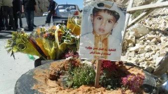 L'image montre un mémorial pour un enfant, avec une photo de lui affichée sur un panneau en bois planté dans un pneu. Autour du panneau, il y a des fleurs multicolores et de la terre. En arrière-plan, on peut voir des personnes rassemblées et un paysage de pierre. Cela semble être un hommage émouvant à un enfant décédé.