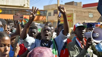 L'image montre une foule de manifestants lors d'une manifestation. Ils semblent exprimer leur colère ou leur détermination, avec des poings levés et des expressions intenses. Certaines personnes tiennent des téléphones portables, tandis qu'une autre utilise un mégaphone pour s'exprimer. En arrière-plan, on aperçoit des bâtiments et des drapeaux, ce qui donne une idée d'un contexte de protestation pour une cause déterminée. L'atmosphère semble énergique et passionnée.