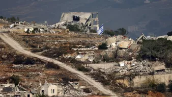 L'image montre un paysage de ruines, probablement dans une région touchée par un conflit. À l'arrière-plan, on peut voir des bâtiments détruits, avec des débris éparpillés. Une route de terre serpente à travers le paysage, et un drapeau israélien apparaît au premier plan, signalant la présence israélienne dans la zone. Le paysage est montagneux, avec des collines en arrière-plan, suggérant un environnement rural. L'image évoque une atmosphère de désolation et de conflit.