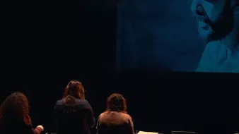 L'image montre un groupe de personnes de dos, se tenant devant une projection qui affiche une image en noir et blanc d'un homme portant un chapeau. Les personnes semblent engagées dans une activité, peut-être en lien avec la projection. Au premier plan, il y a un bureau avec un ordinateur et d'autres objets. L'éclairage crée une atmosphère intrigante autour de la scène.