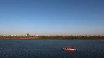 L'image montre une scène paisible d'un cours d'eau avec un ciel dégagé et sans nuages. Au centre, on peut voir un petit bateau naviguant sur les eaux calmes. À l'arrière-plan, on distingue une ligne d'horizon urbaine, avec des bâtiments et des arbres le long de la rive. L'ambiance générale est tranquille et ensoleillée, suggérant une belle journée.