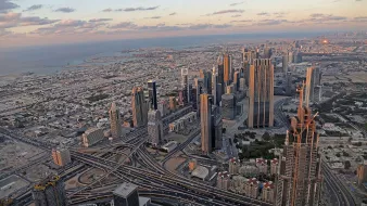L'image montre un panorama impressionnant d'une ville moderne, probablement Dubai. On y voit de nombreux gratte-ciels élégants s'élevant vers le ciel, ainsi qu'un réseau complexe de routes et d'autoroutes serpentant à travers la métropole. À l'arrière-plan, on aperçoit la mer et des paysages urbains qui s'étendent à perte de vue. Le ciel présente des teintes douces de bleu et d'orange, témoignant d'un coucher de soleil, ce qui ajoute une ambiance tranquille à la scène urbaine animée.