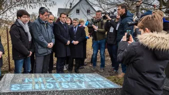 L'image montre un groupe de personnes rassemblées autour d'une stèle ou d'une tombe. Parmi elles, on peut distinguer des individus en tenue formelle, semblant assister à une cérémonie hommage. Un enfant tient un téléphone pour filmer ou prendre une photo. Des journalistes, avec des caméras et des micros, sont également présents, suggérant que l'événement est médiatisé. En arrière-plan, on aperçoit une maison et des arbres, indiquant un cadre extérieur.
